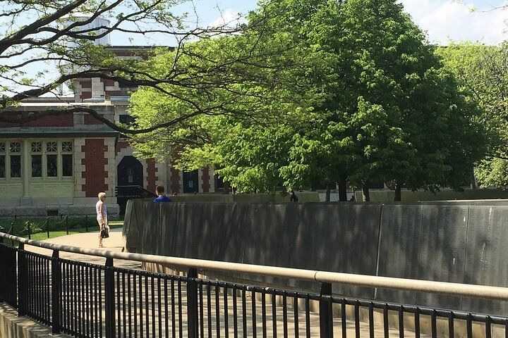 A person walks on a path near a concrete wall, trees, and a brick building.