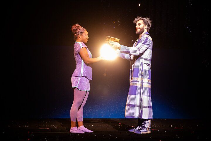Two people, one in purple sportswear and one in a checkered robe, hold a glowing orb on a dark stage.