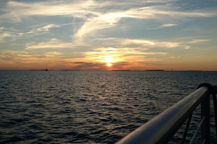 Sunset over the ocean, with a metal railing in the foreground. Orange and yellow hues in the sky.