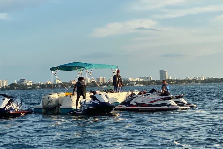 Jet skis near a pontoon boat in the water. People on boat and jetskis; city skyline in the background.