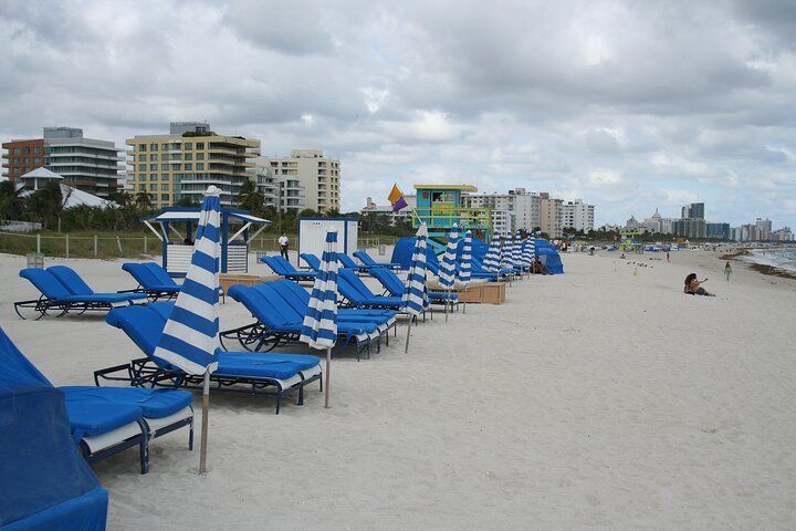 Beach with blue lounge chairs and umbrellas, buildings in the background. Overcast day.