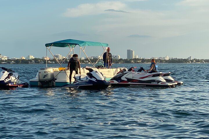 Jet skis and people near a white pontoon boat on the water, city skyline in the background.