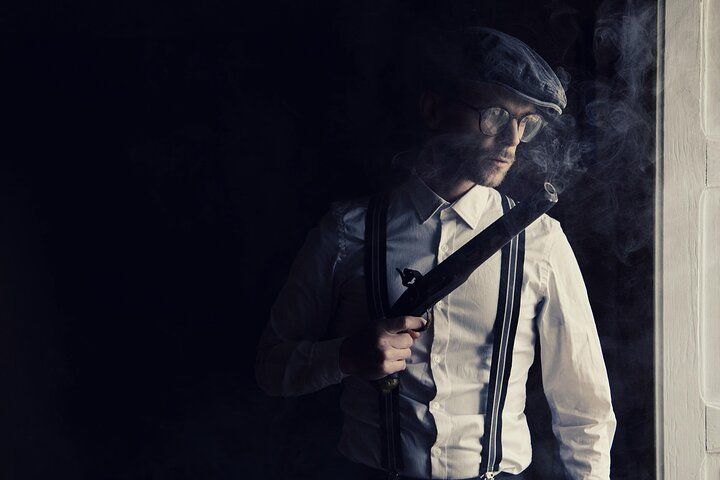 Man in flat cap, holding object, smoking, looking out a window.