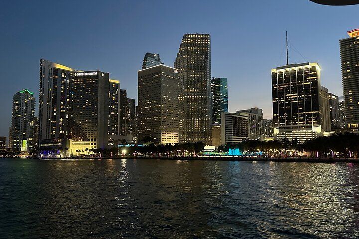 Nighttime view of Miami skyline with illuminated buildings reflecting on water.
