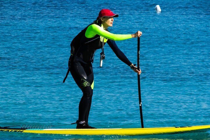 Woman in wetsuit paddle boarding on blue water, wearing red cap, holding paddle.