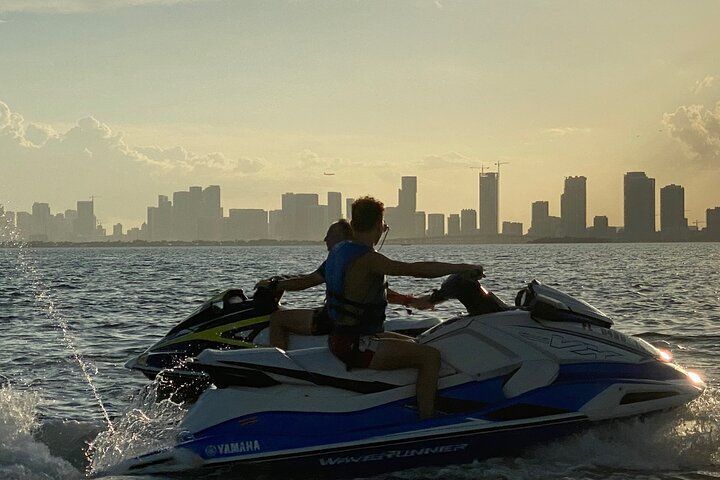 Two people on jet skis on water, city skyline in the background.