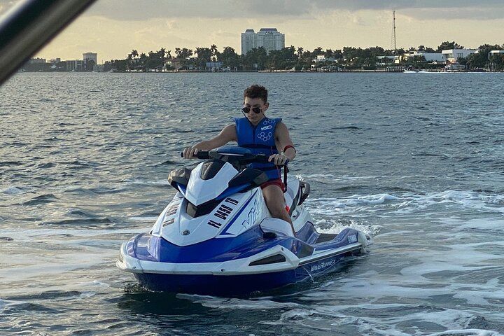 Teenager on blue and white jet ski on water, wearing sunglasses and life vest. Buildings in background.