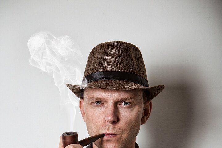 Man in fedora and suit, smoking a pipe, exhaling smoke.