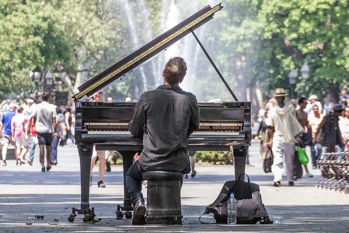 Pianist plays a grand piano outdoors in a park with a fountain, surrounded by people walking.