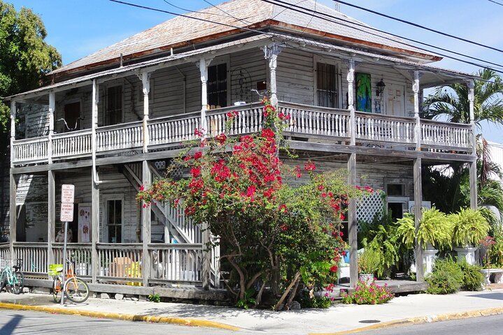 Two-story wooden house with wrap-around porches, red flowers, and a bicycle on the sidewalk.