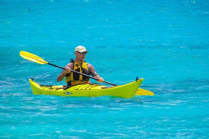 Man in a yellow kayak paddles on turquoise water, wearing a life vest and hat.