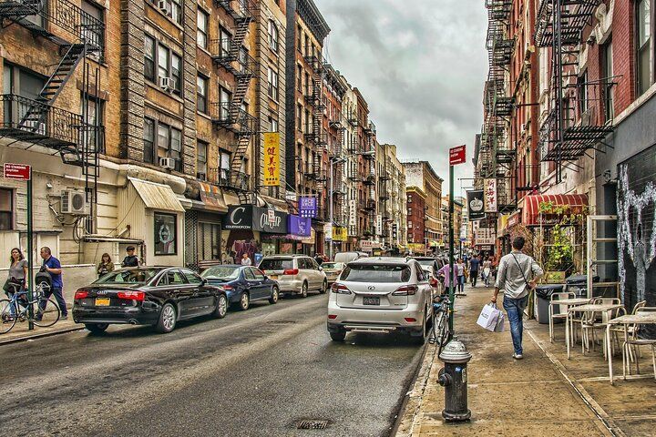 Street scene: Cars and pedestrians on a city street lined with brick buildings, fire escapes, shops, and a cloudy sky.