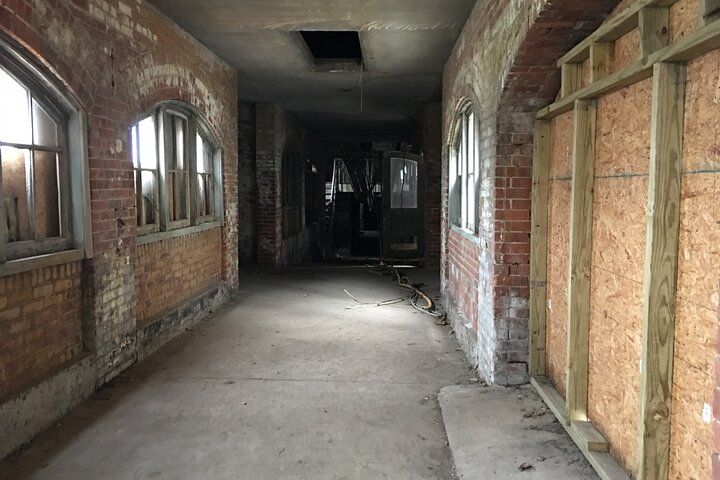 Dilapidated brick hallway with arched windows, boarded walls, and a dark opening at the end.