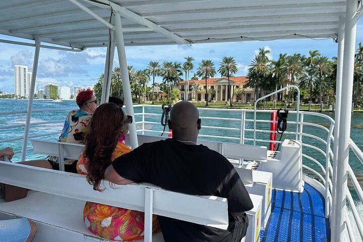 People on a boat viewing waterfront houses. Blue water, sunny sky. Palm trees line the shore.