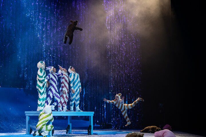 Performers in striped costumes watch a bear puppet flying in a theatrical setting with falling sparkle effects.