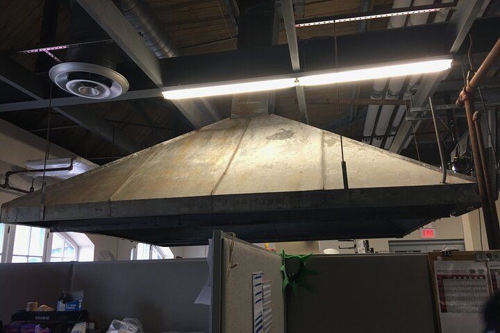 Large, square, metal exhaust hood suspended from a ceiling in an office setting, above cubicle walls.