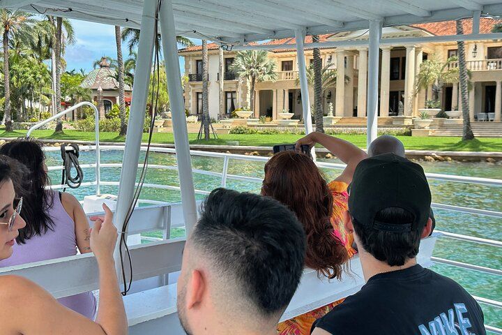 People on a boat viewing a large mansion with columns, palm trees, and clear water.