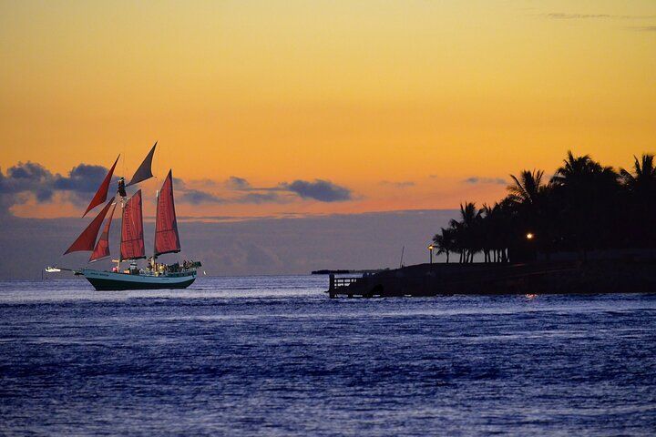 Sailboat with red sails on ocean at sunset, silhouetted palm trees and colorful sky.