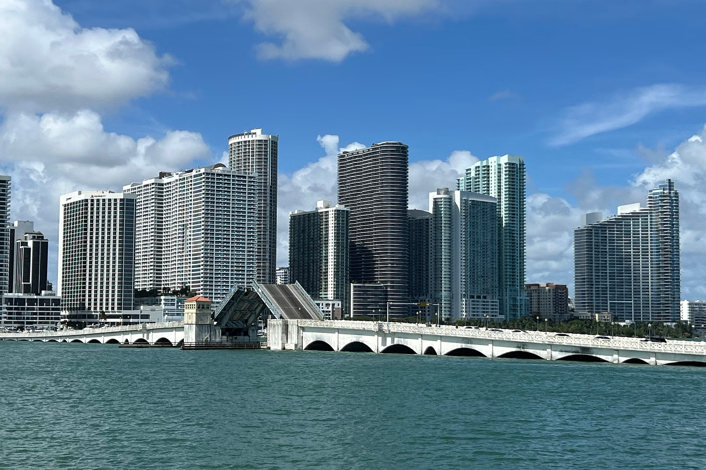 Miami skyline with a bridge over water, blue sky, and puffy clouds.