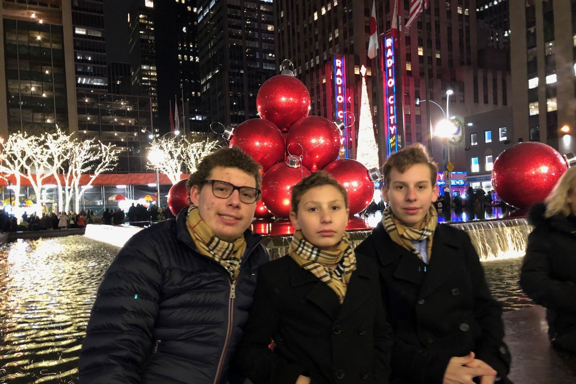 Three people pose in front of red balloons and Rockefeller Center at night.
