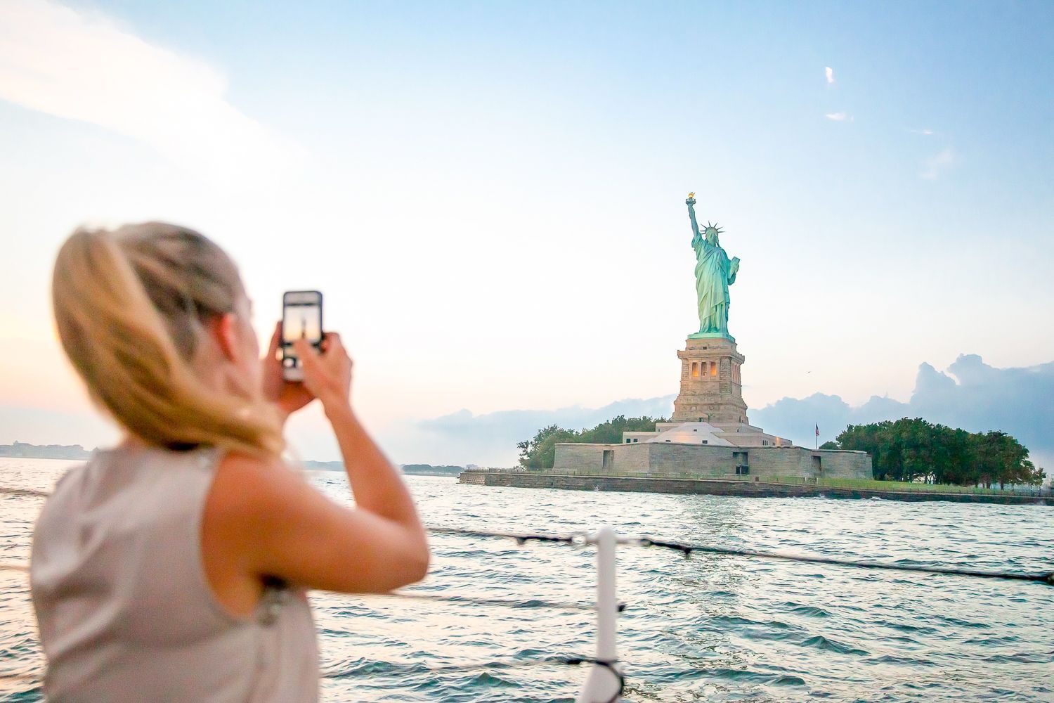 Woman taking a photo of the Statue of Liberty from a boat on the water; sunset.