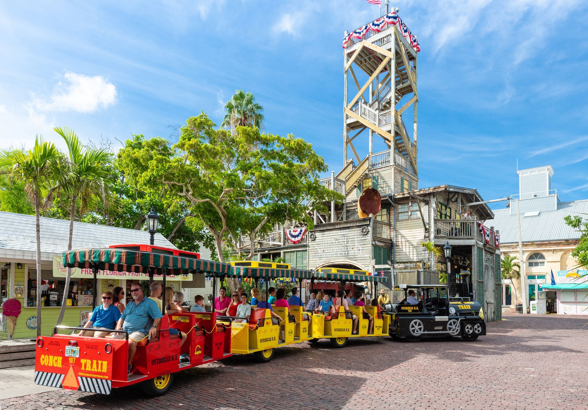 Red train filled with tourists near a weathered building with a tall tower in Key West.