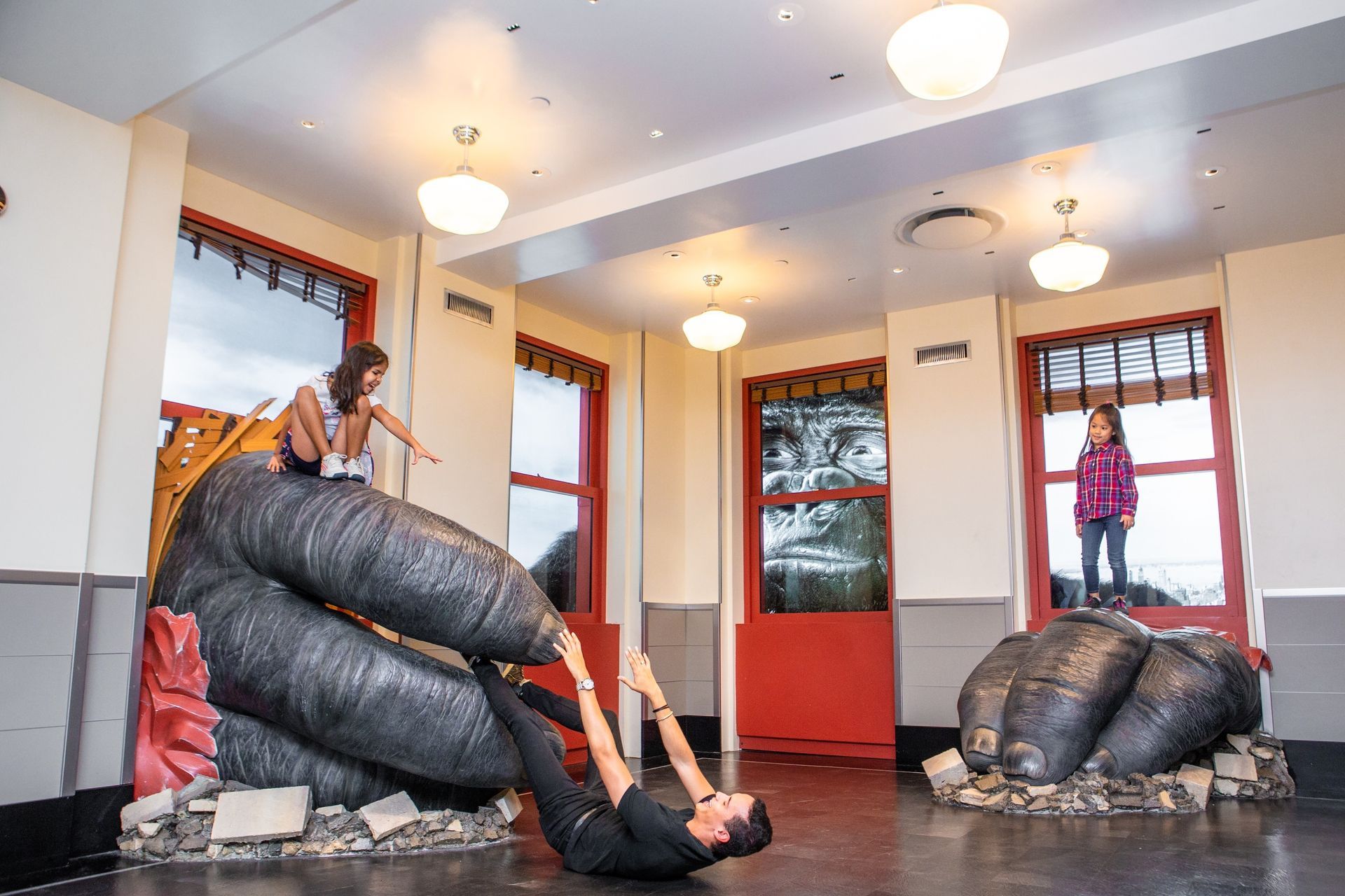 People interacting with a King Kong hand sculpture inside a building, with one person hanging from its fingers.