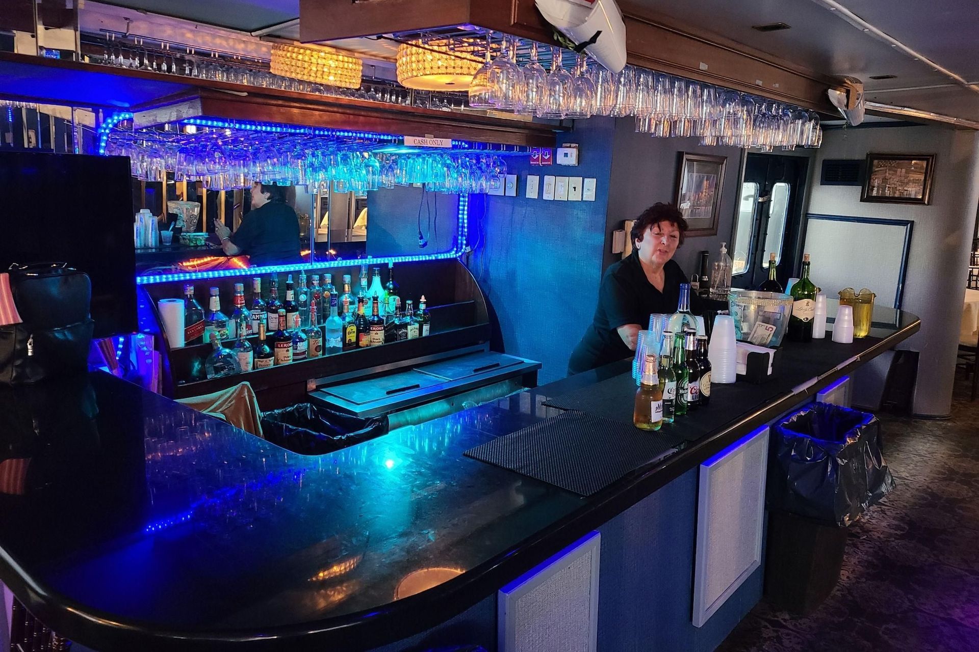 Bar interior with a bartender standing behind the counter, bottles and glasses. Blue lights are illuminating the bar.