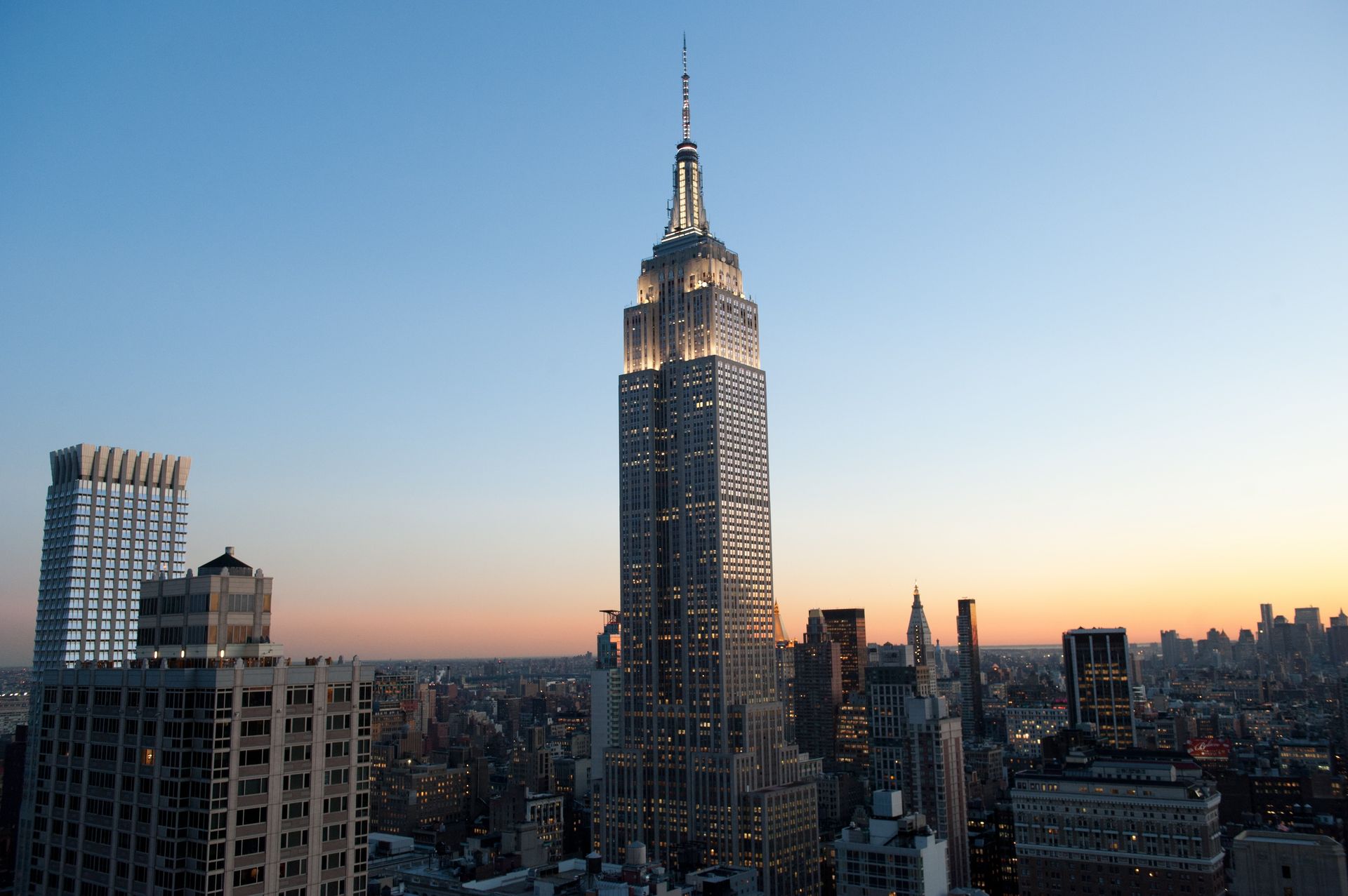 Empire State Building, New York City skyline at dusk. Tower lit, surrounded by other buildings, blue sky.