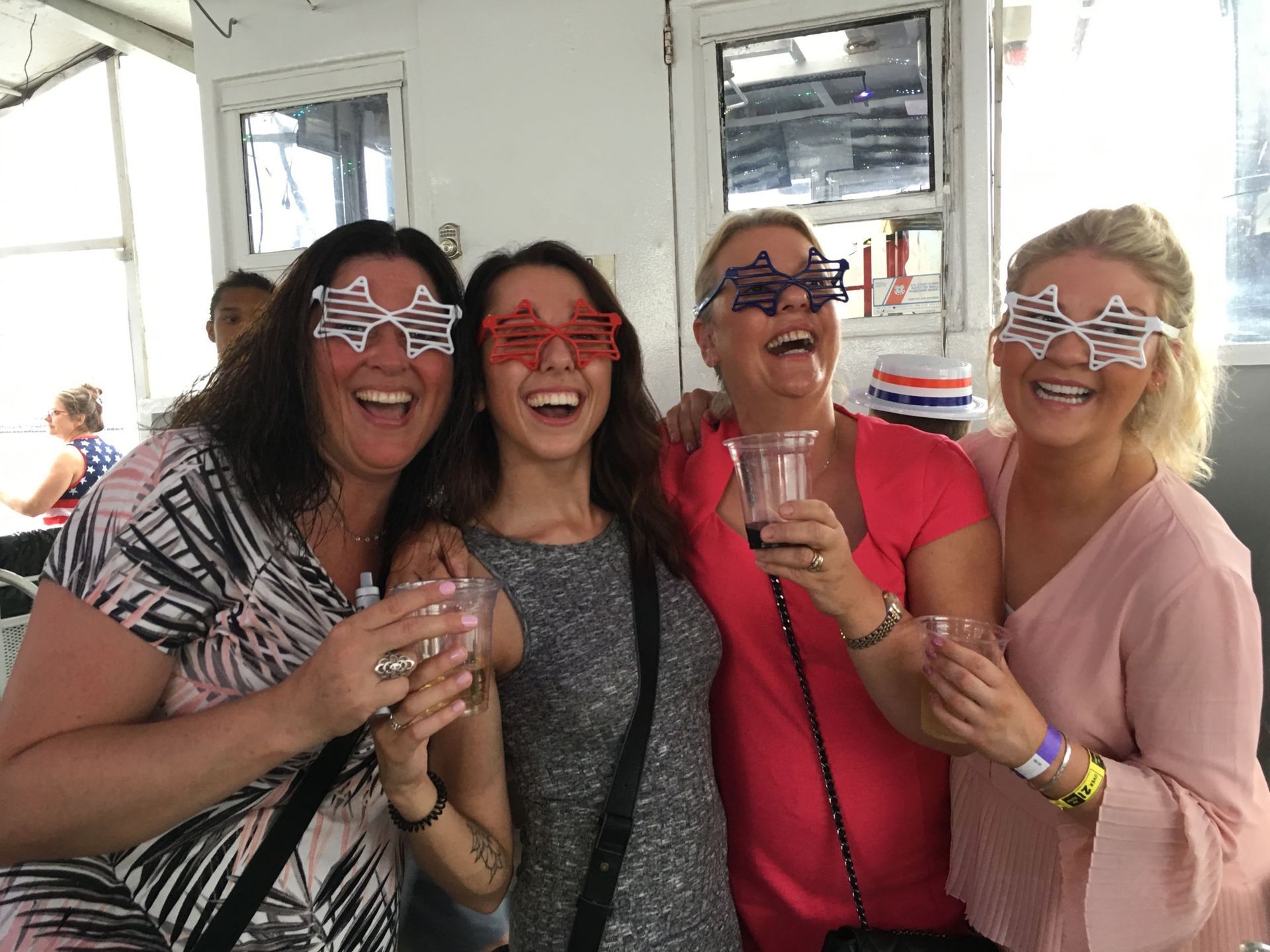 Four women laughing, wearing star-shaped glasses. Holding drinks, in a brightly lit room.