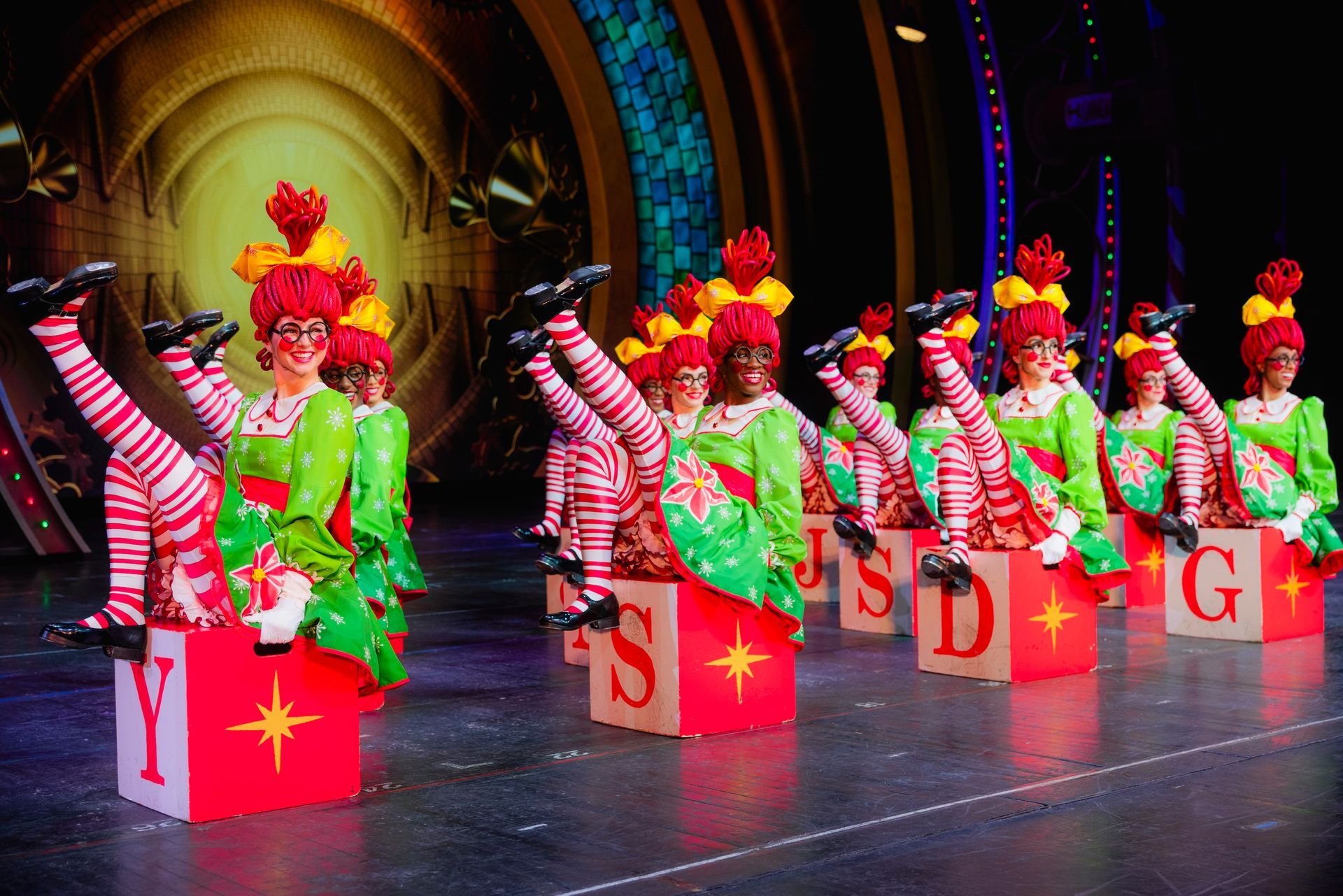 Dancers in elf costumes with striped legs and red and green outfits, sitting on lettered boxes, raising legs. Stage setting.