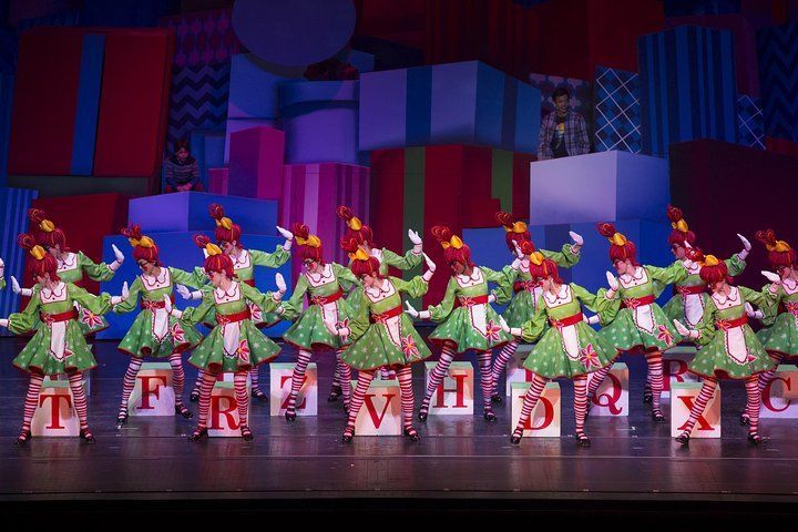 Dancers in green costumes with red trim perform on stage with gift box backdrop.