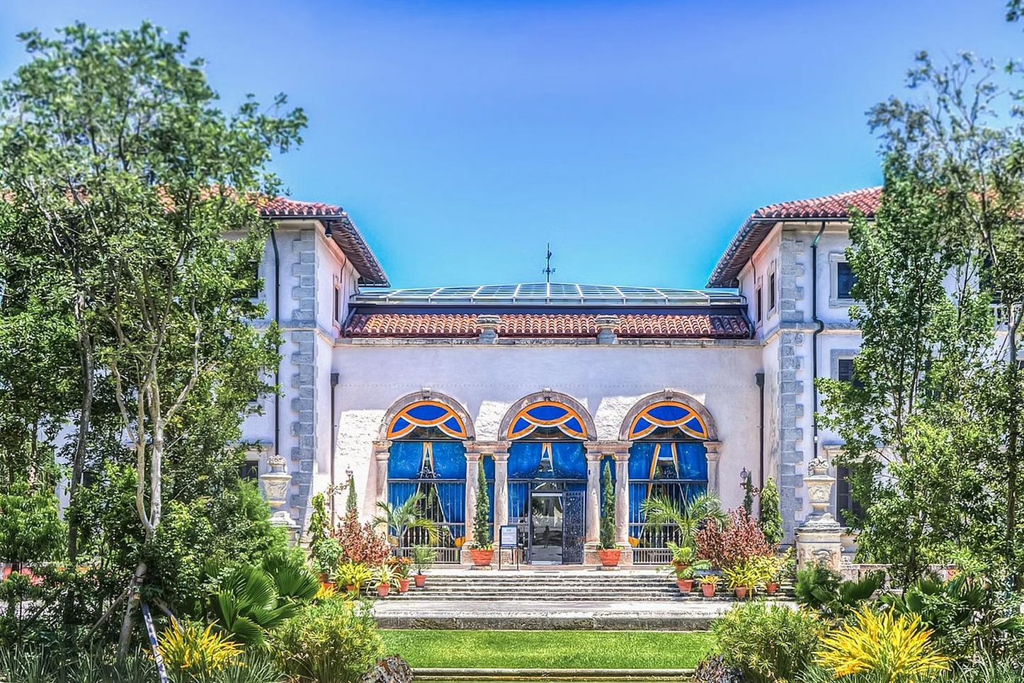 Ornate mansion with blue accents, arched windows, and lush greenery under a clear blue sky.