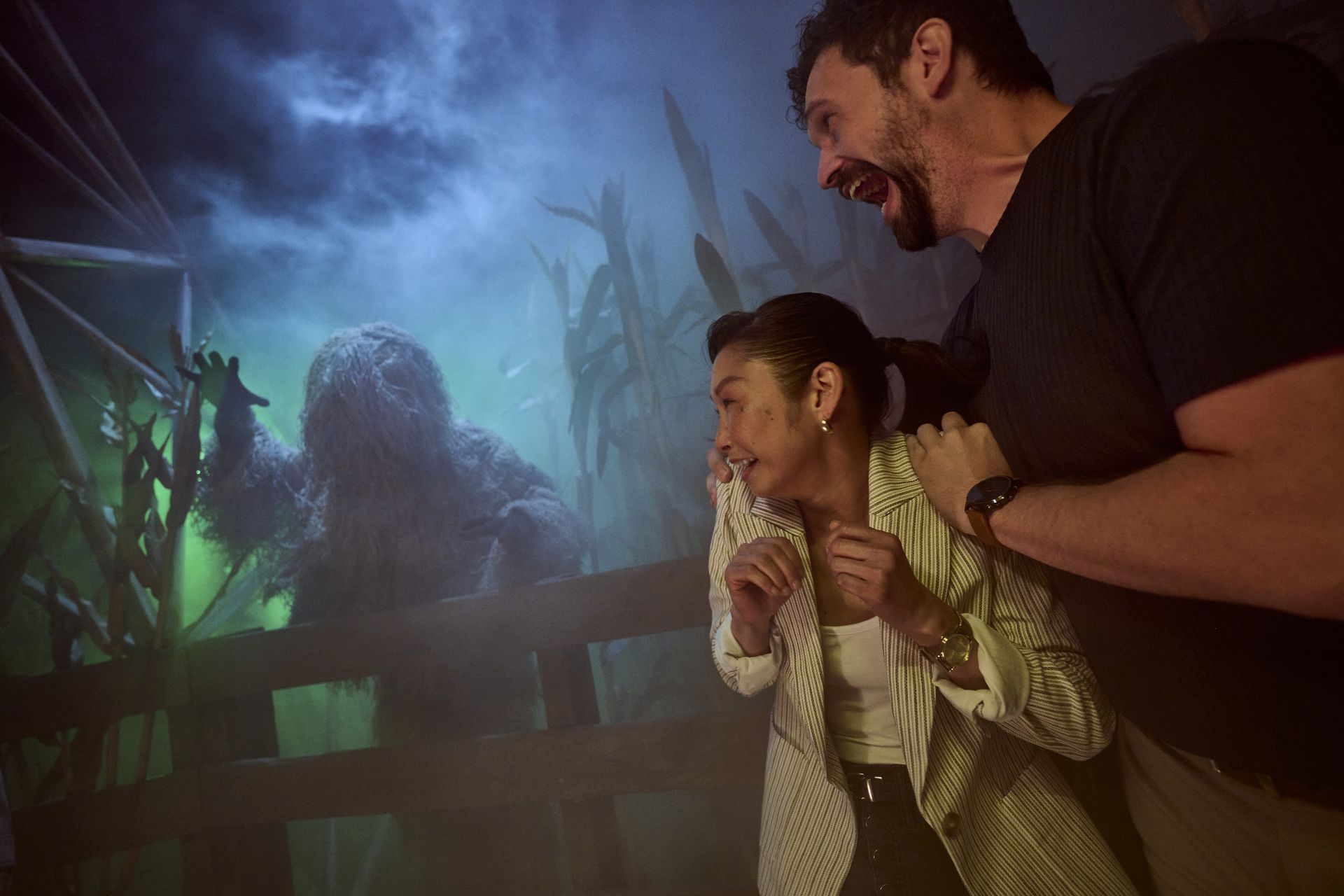 Two people react in fright as a swamp creature lunges at them in a foggy haunted house setting.