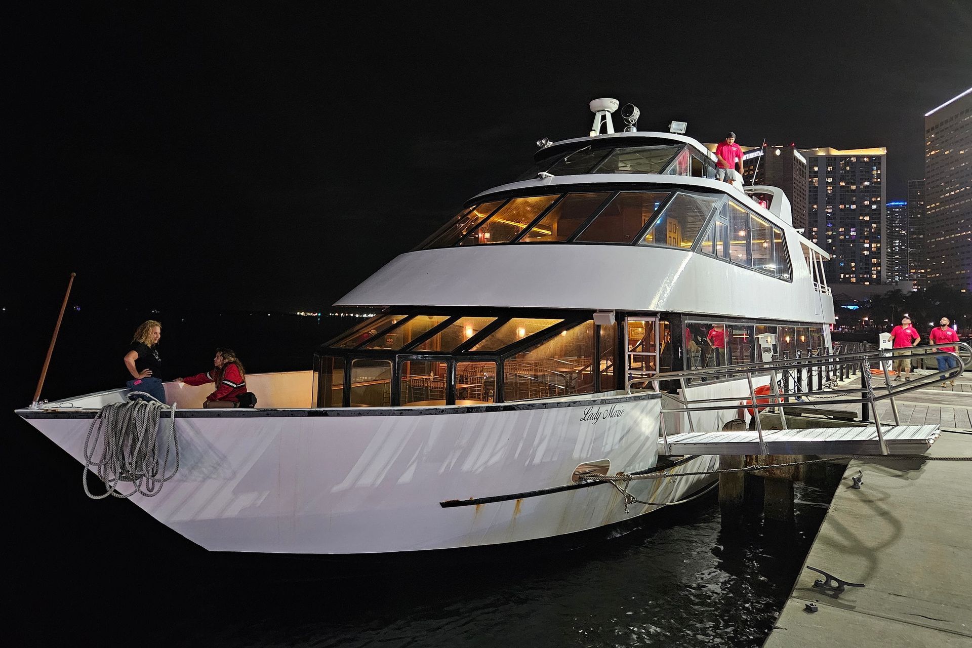 A white yacht docked at night with people visible on the deck and a city skyline in the background.