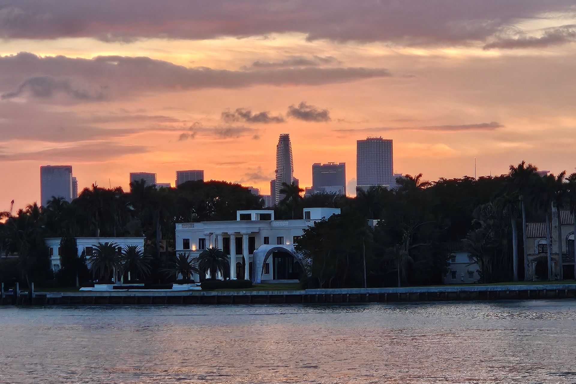 Skyline at sunset, with buildings silhouetted behind palm trees and a body of water.