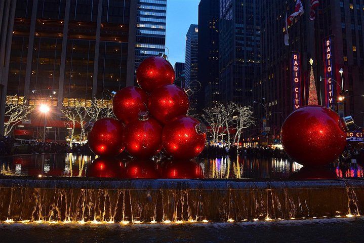 Red Christmas ornaments in a fountain, with city buildings in the background at dusk.