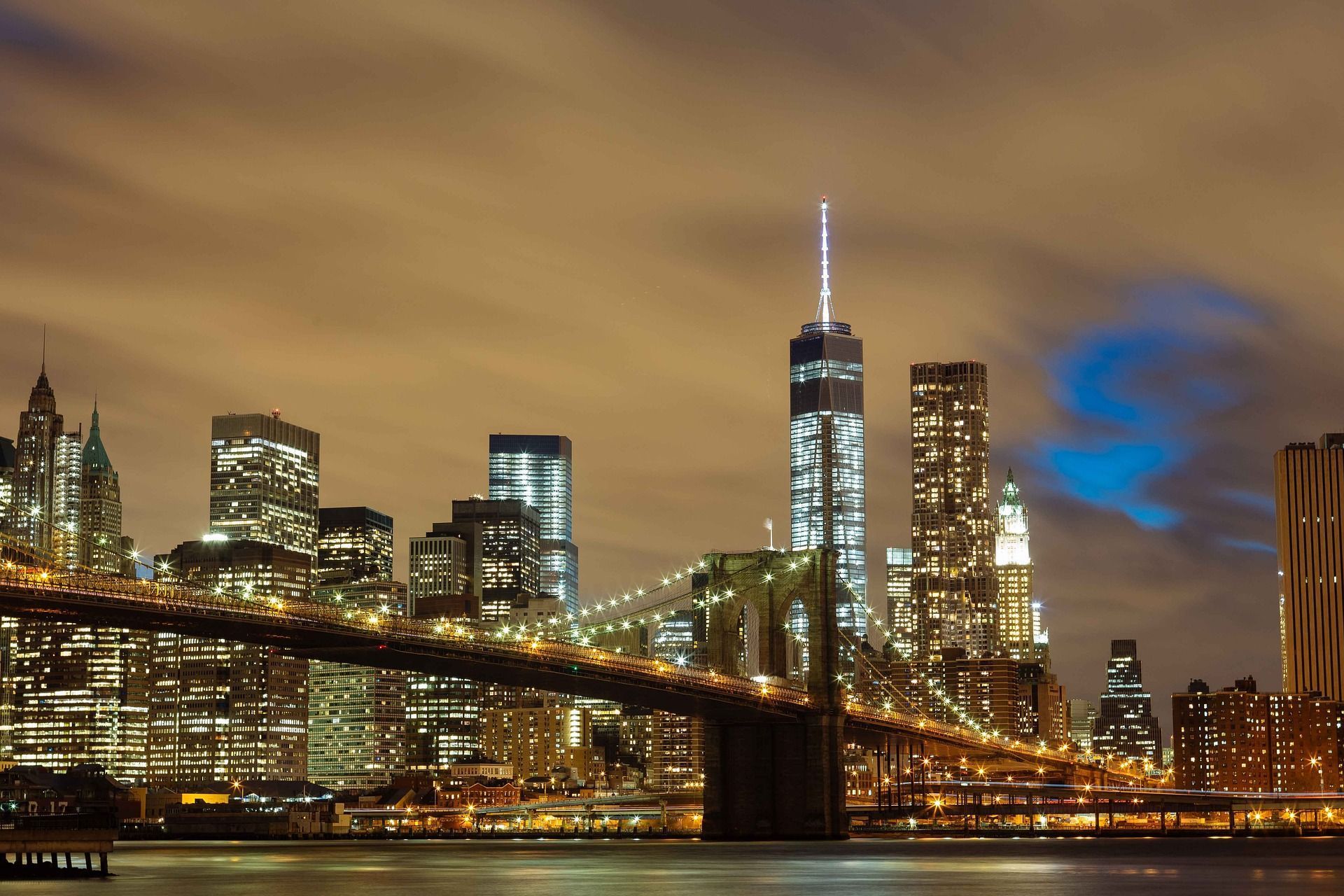 New York City skyline at night with Brooklyn Bridge. Illuminated buildings and a cloudy sky.