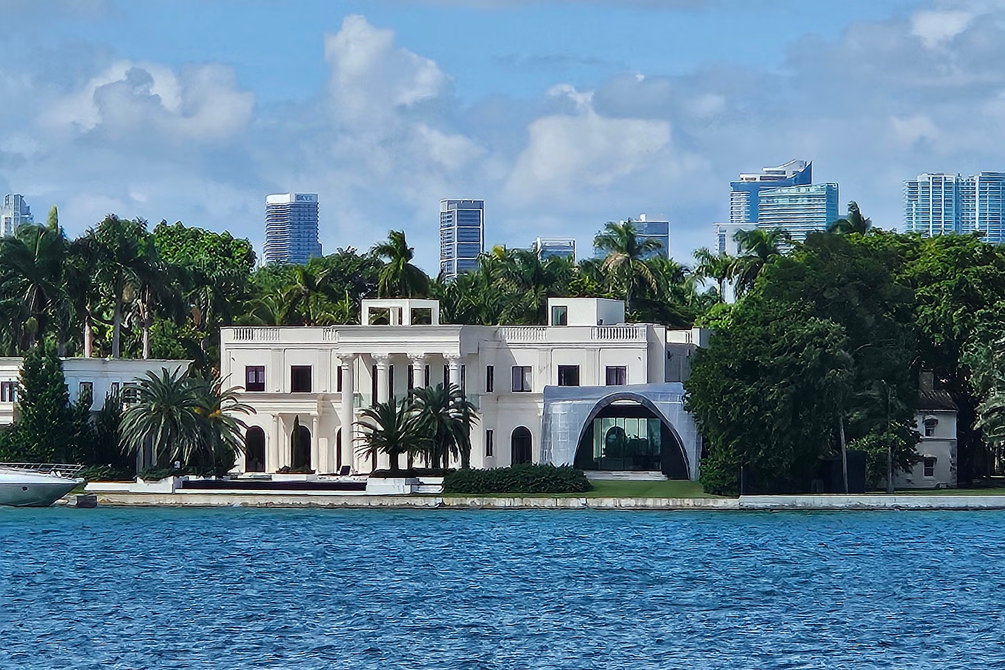 Large white mansion by blue water, with a skyline and green trees in the background.