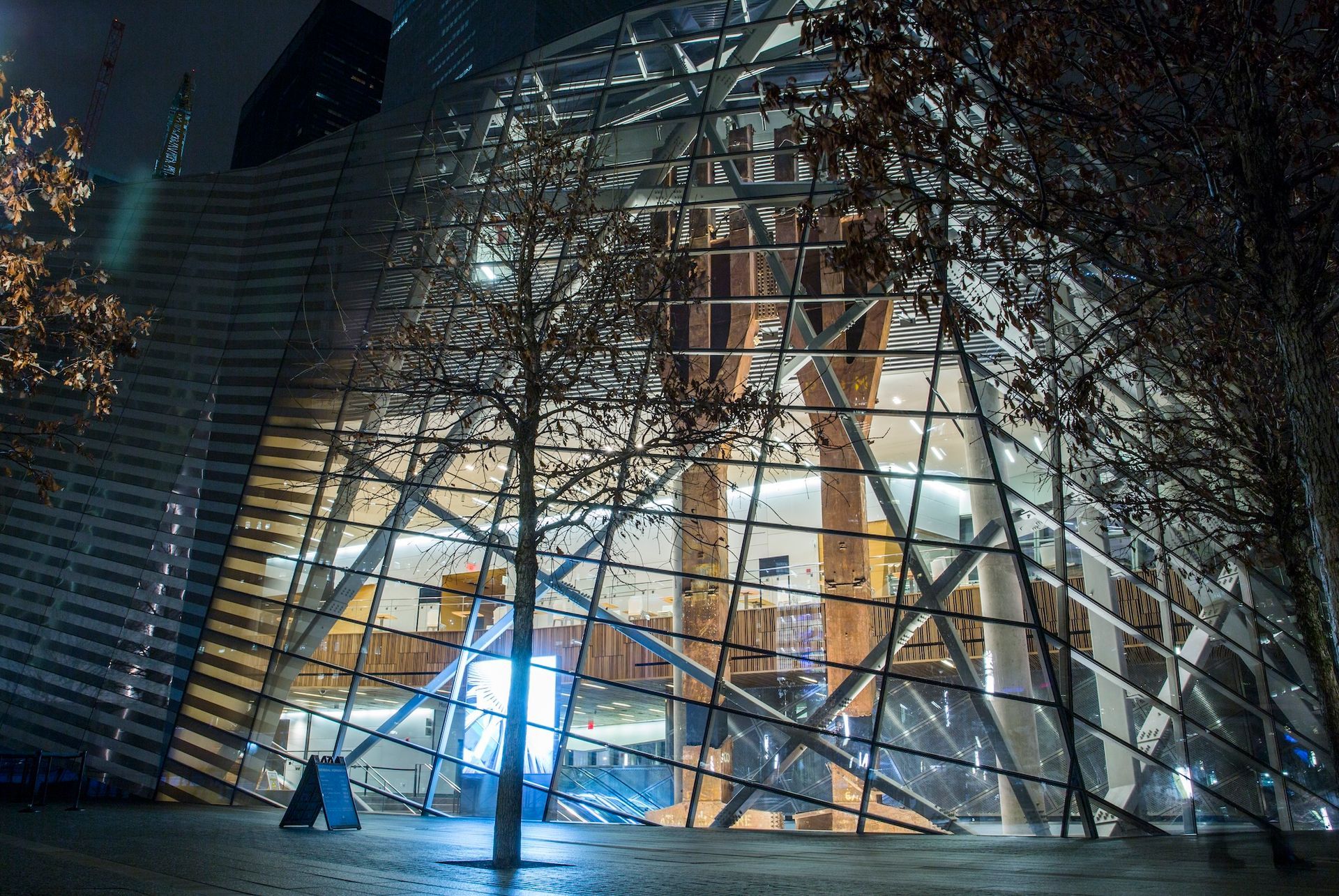 Modern glass building at night, trees in foreground, lights glow inside.