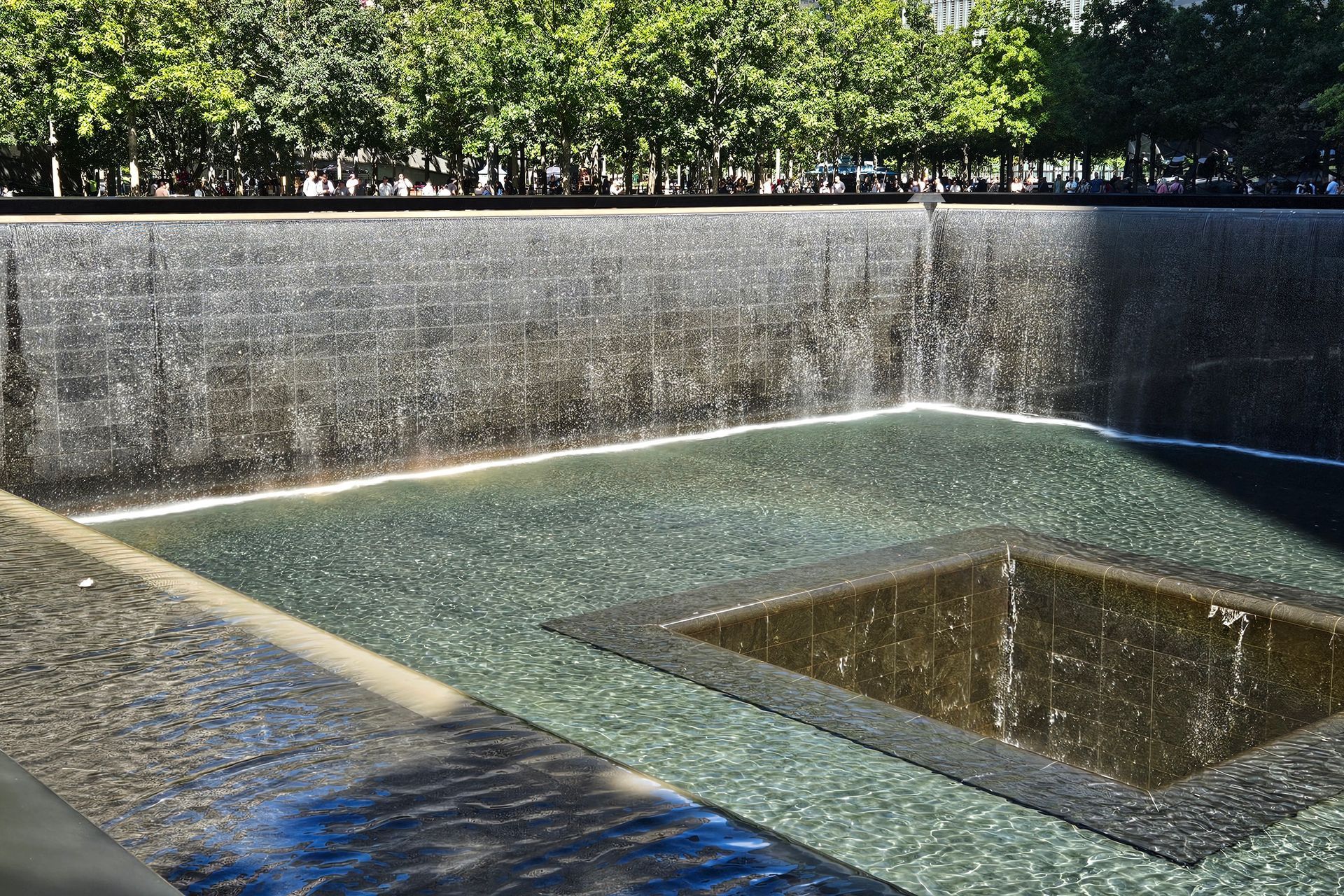 9/11 Memorial reflecting pool with cascading water into a square basin. Sunlight creates sparkling reflections.