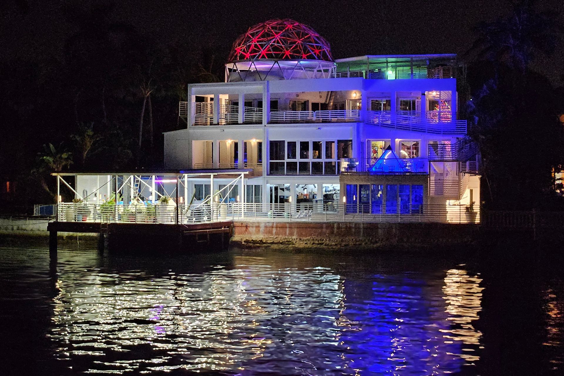 Night view of a multi-story building with a glowing red dome and blue lighting reflected in water.