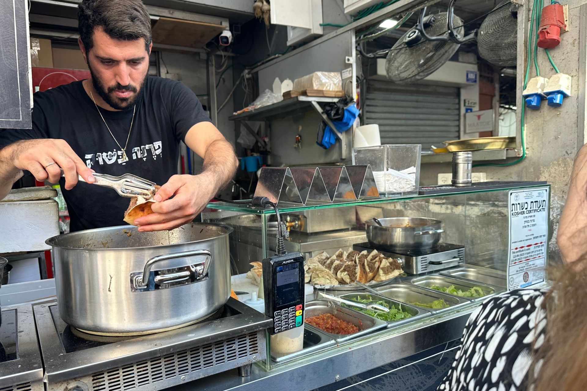 Man preparing food in a small kitchen with pots, ingredients, and customer visible.