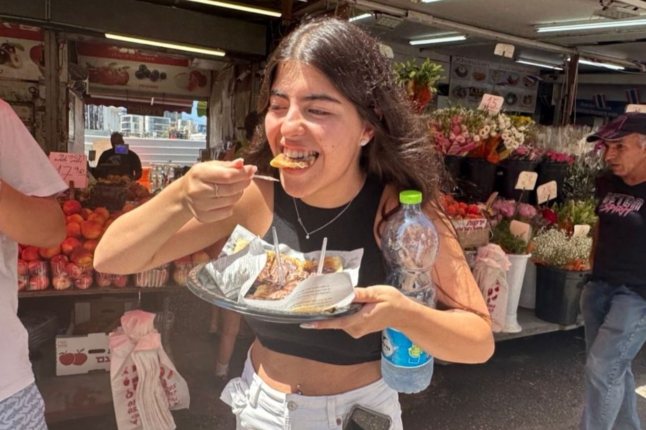 Woman eating from a plate outdoors. She’s smiling with a fork in hand, near a flower stand and food vendors.
