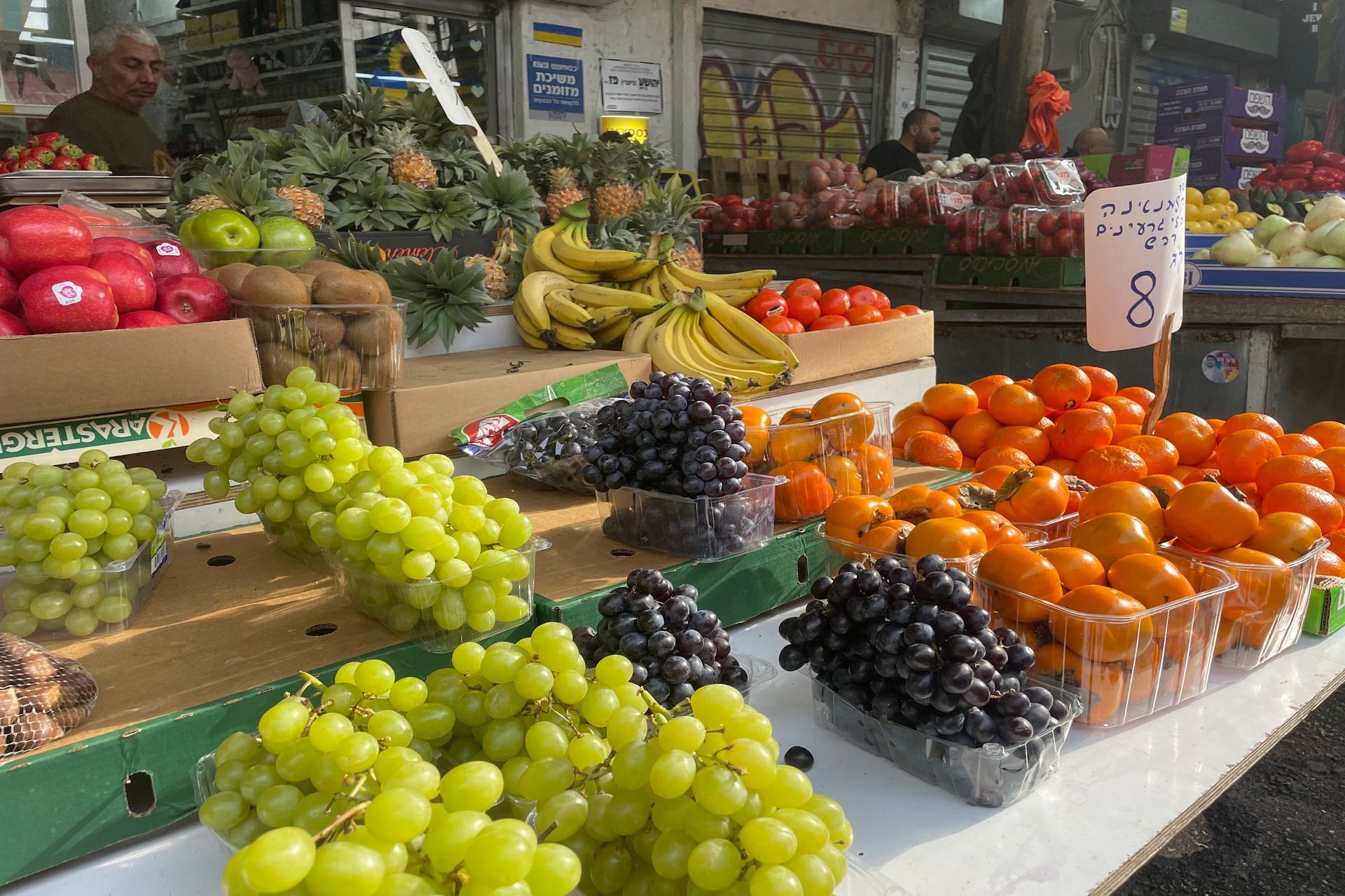 Fruit stand with grapes, oranges, bananas, and other produce on display; shoppers and vendors in the background.