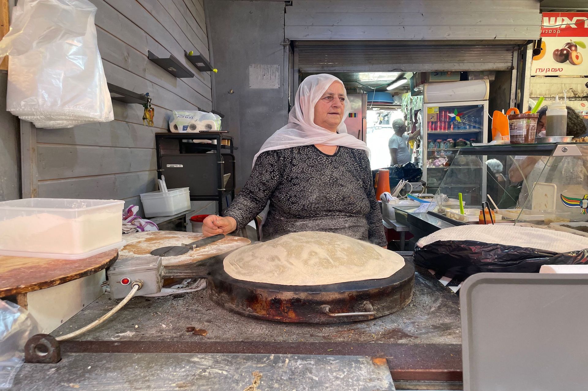 Woman in headscarf prepares food at an outdoor stall, with a large, flat bread-like item in front of her.