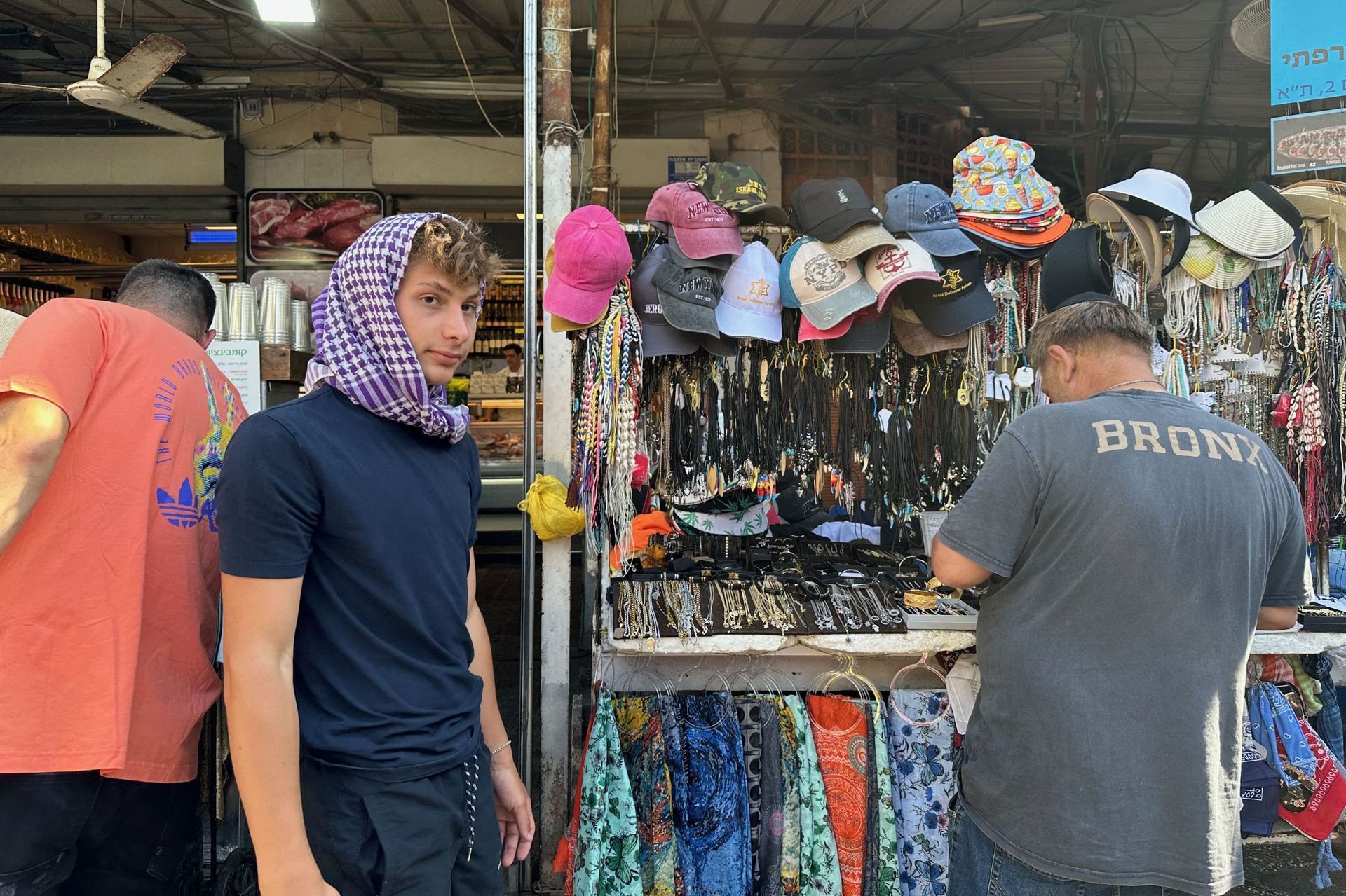 A young man with a headscarf stands near a shop displaying hats and jewelry.