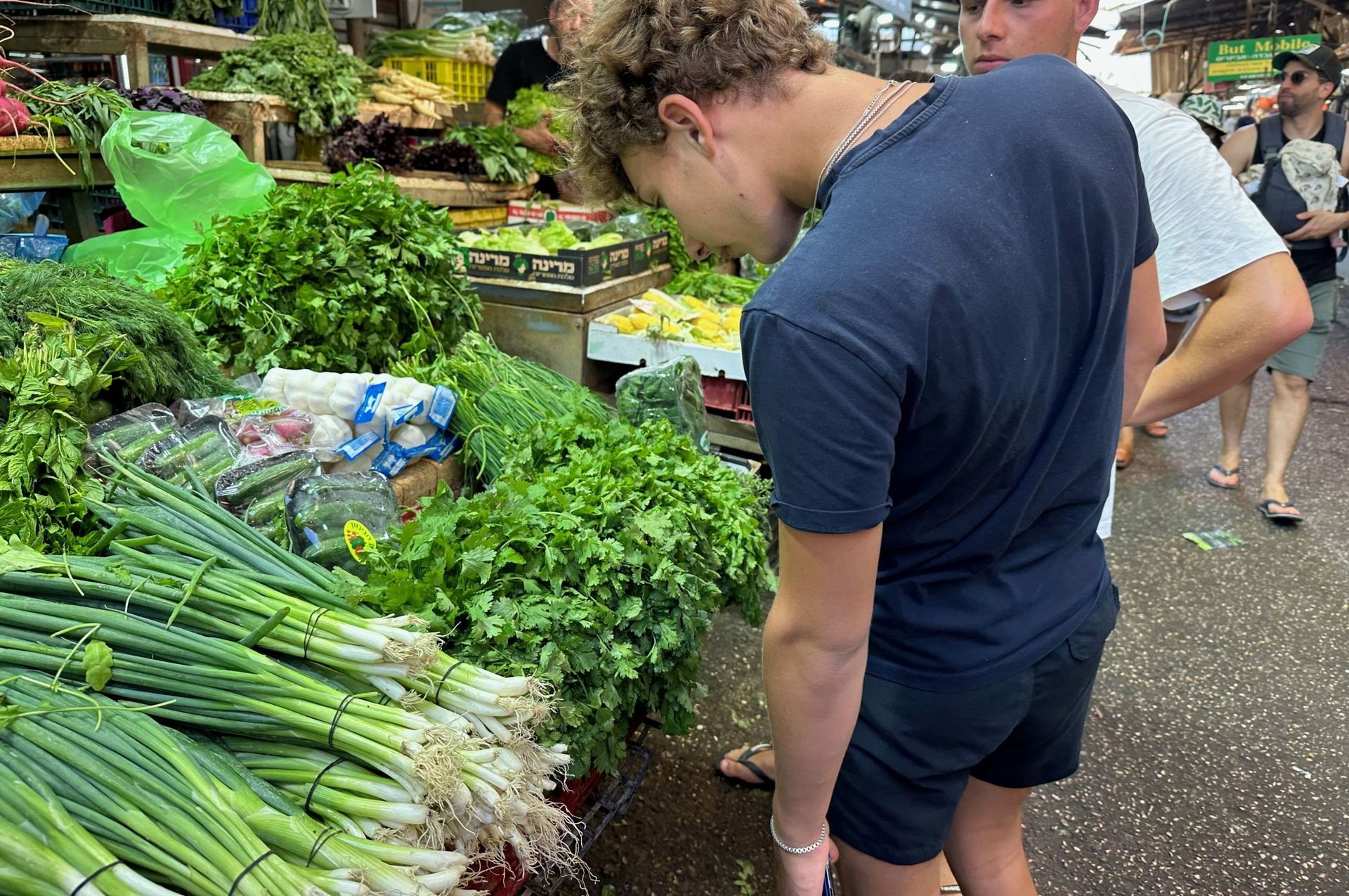 Man at a farmer's market inspecting green onions and other fresh produce; other people are nearby.