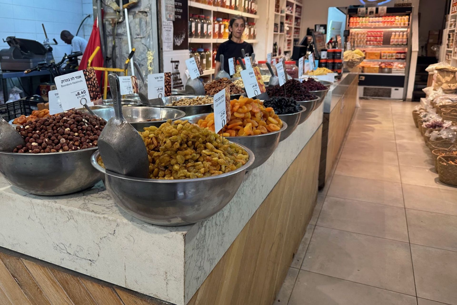 Bowls of dried fruits and nuts in a market, with customers and store workers present.