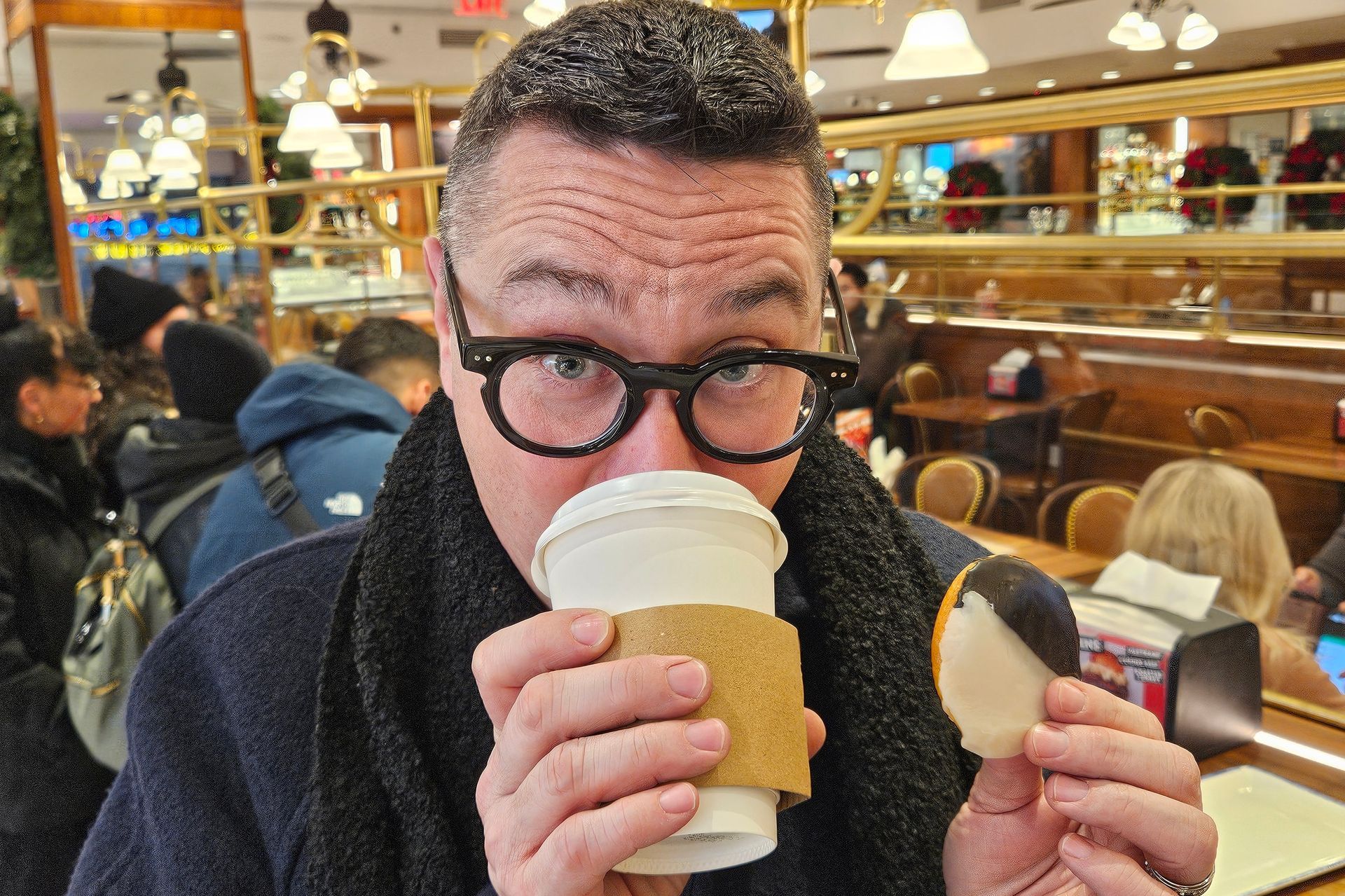Man drinking coffee, holding black-and-white cookie, inside a busy cafe. He's wearing glasses and a scarf.